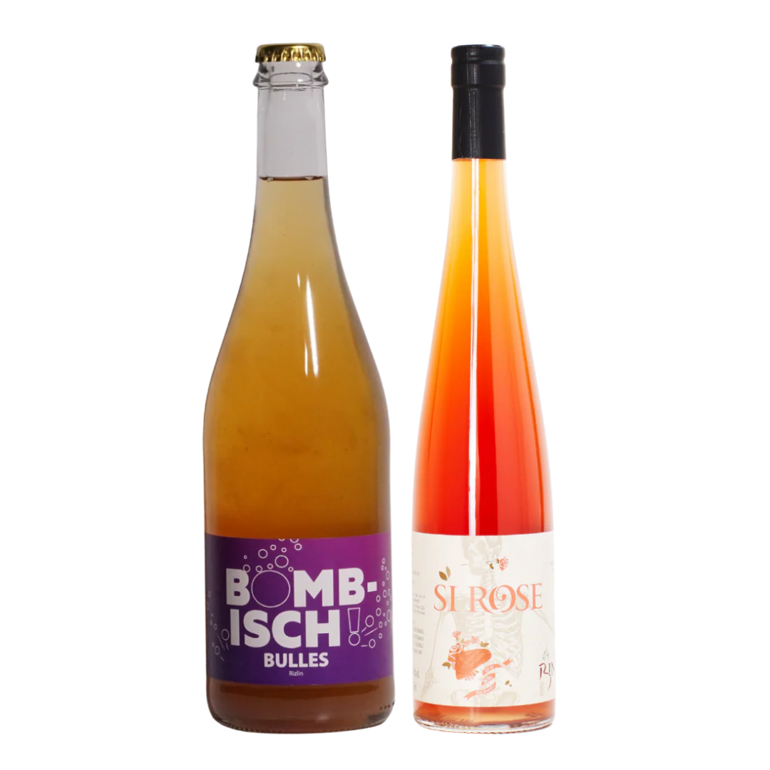 Two beverage bottles on a plain white background: a clear glass bottle of Bombisch Bulles with amber liquid and a purple label, next to a tall clear bottle of Si Rose with an orange-to-pink gradient liquid and a white label.