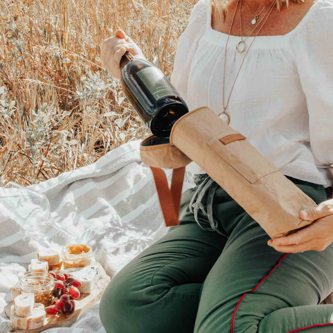 Person sitting outdoors with a picnic setup, getting wine from insulated brown wine tote bag