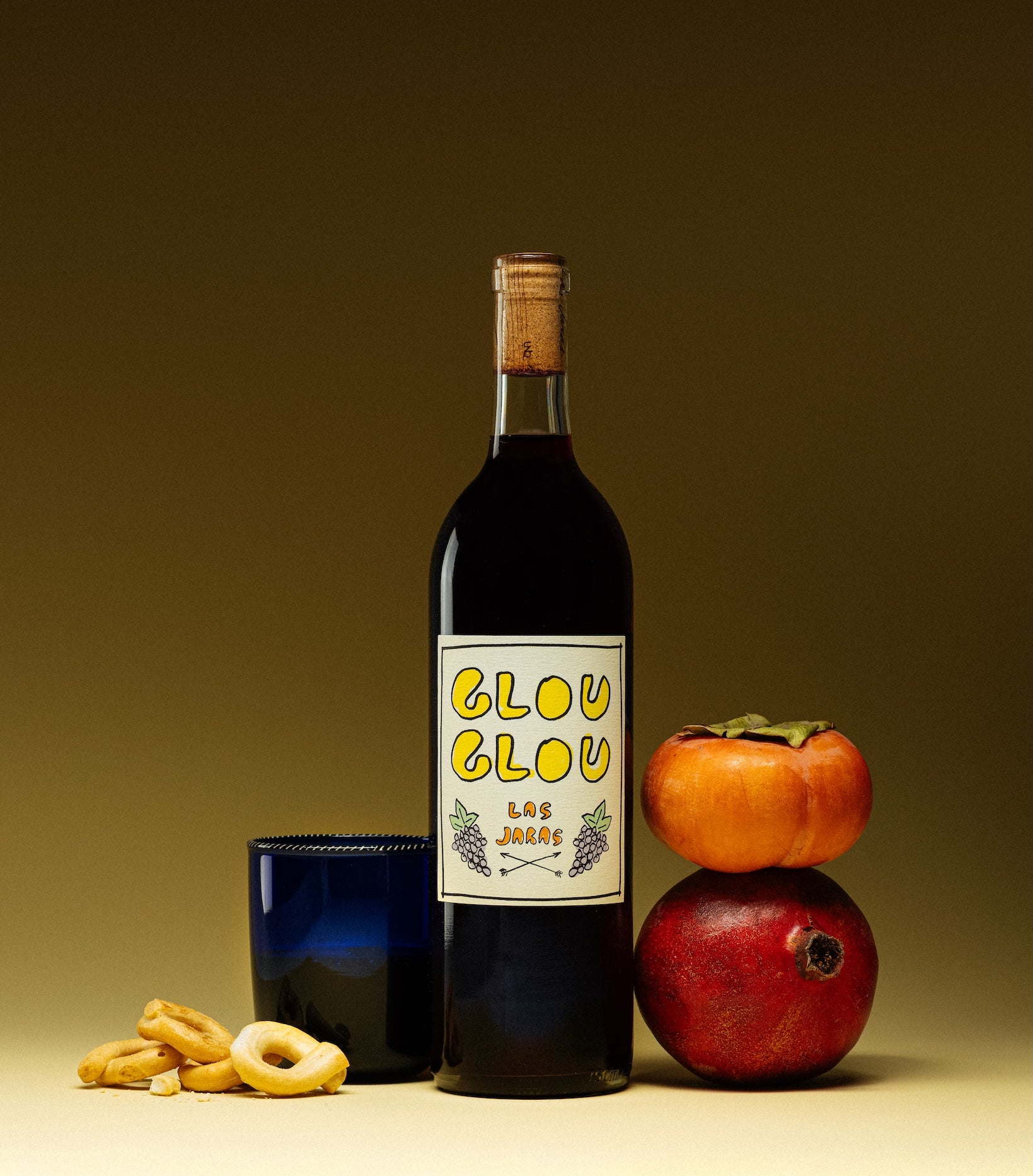 Las Jaras Glou Glou natural red wine bottle with persimmon, pomegranate, and snacks on neutral background for Mysa Natural Wine